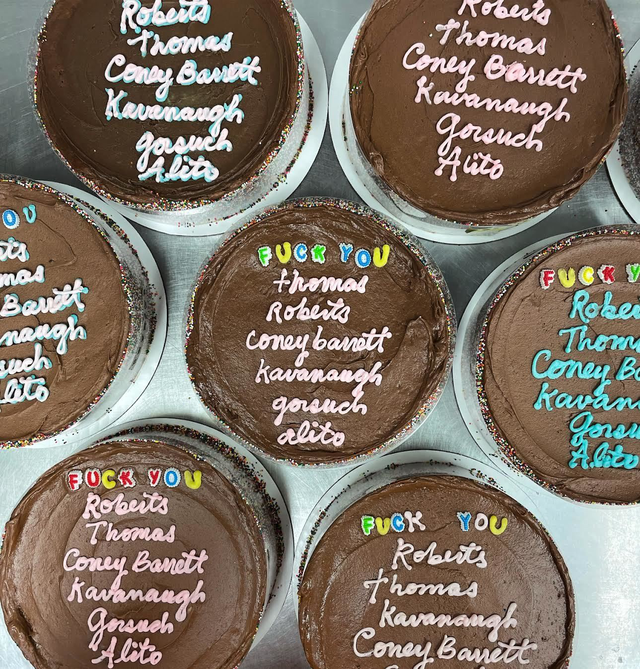 Seven chocolate cakes with rainbow sprinkles on the edges display names of judges and colorful letters spelling “FUCK YOU” above them in various frosting colors.